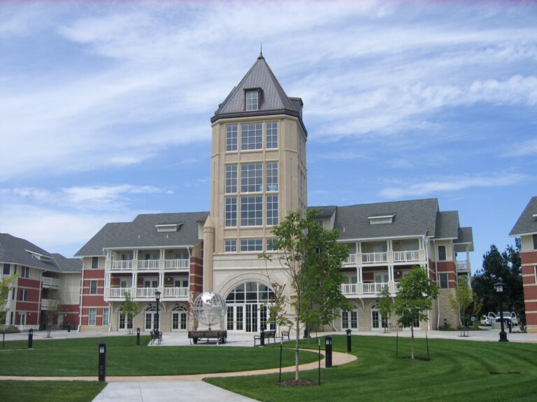 Jardine Apartments complex at Kansas State University, designed by OSE Engineering. The modern residential building features a distinctive cream-colored tower with a peaked metal roof and large windows, flanked by red brick structures with white trim and sloped roofs. A green courtyard with benches and young trees sits in front of the entrance, with the tower's arched base opening onto the plaza below.
